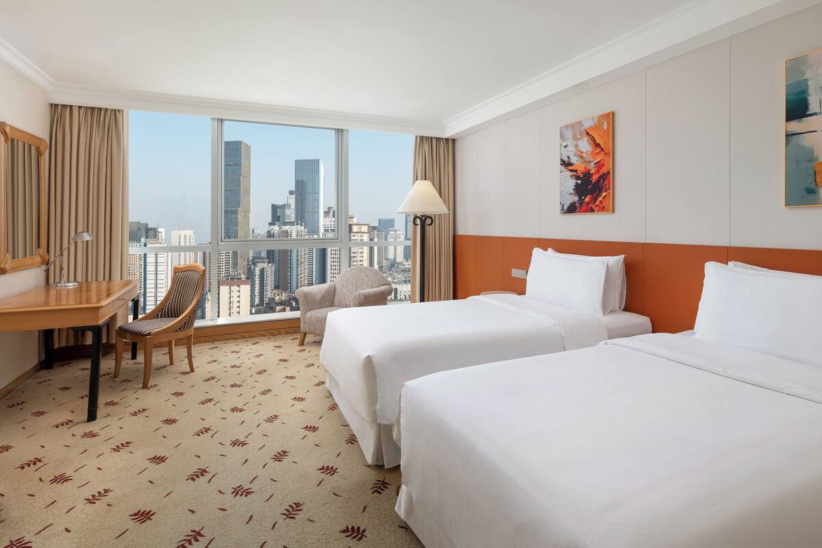 A hotel room with two beds, a desk, a chair, a lamp, and a city view.