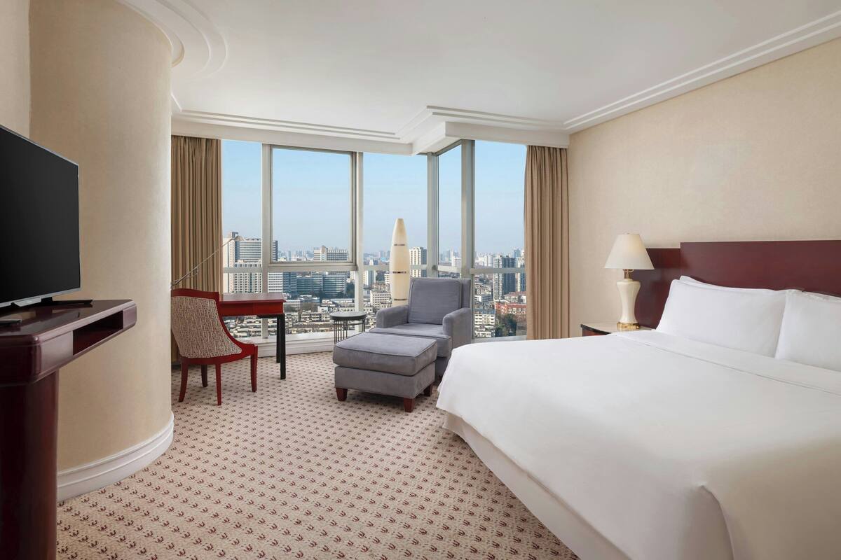 A hotel room with a large bed, a TV, a chair, a small table, and a view of the city.
