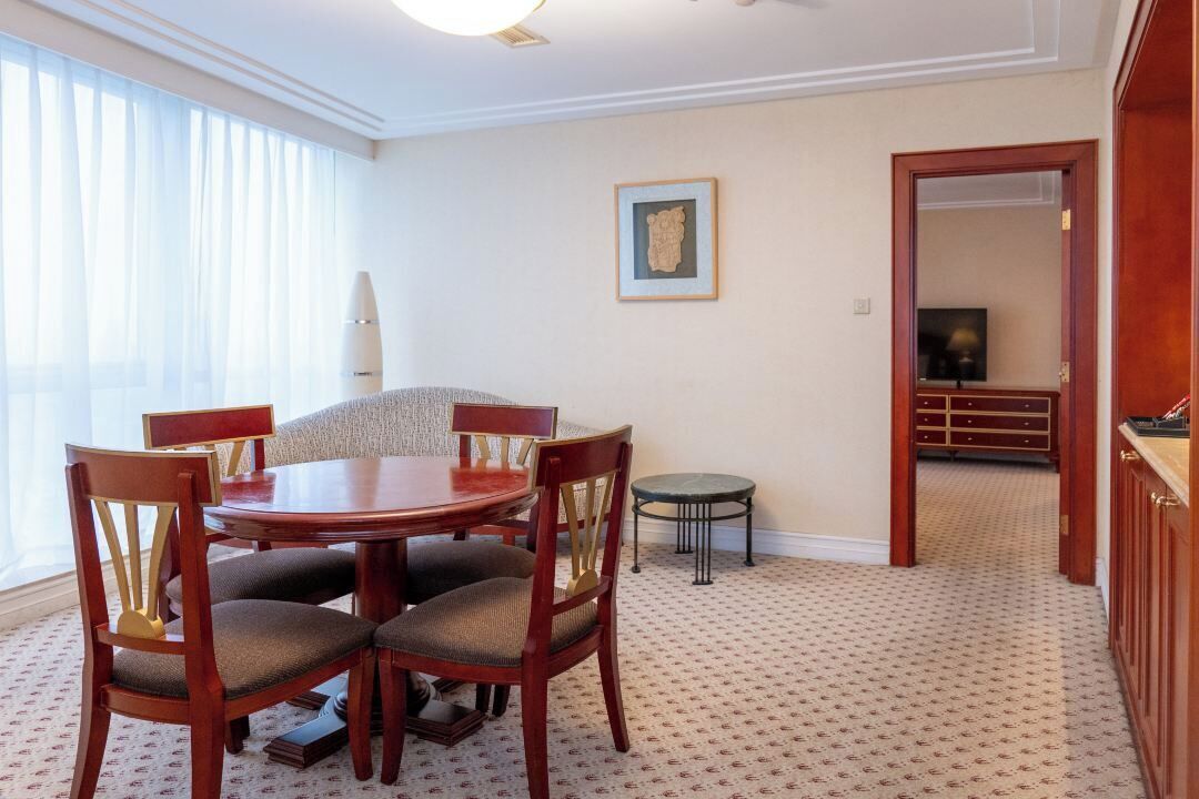 Un comedor con una mesa redonda, cuatro sillas, una mesita auxiliar, un cuadro enmarcado en la pared y vistas a otra habitación a través de un vano abierto.