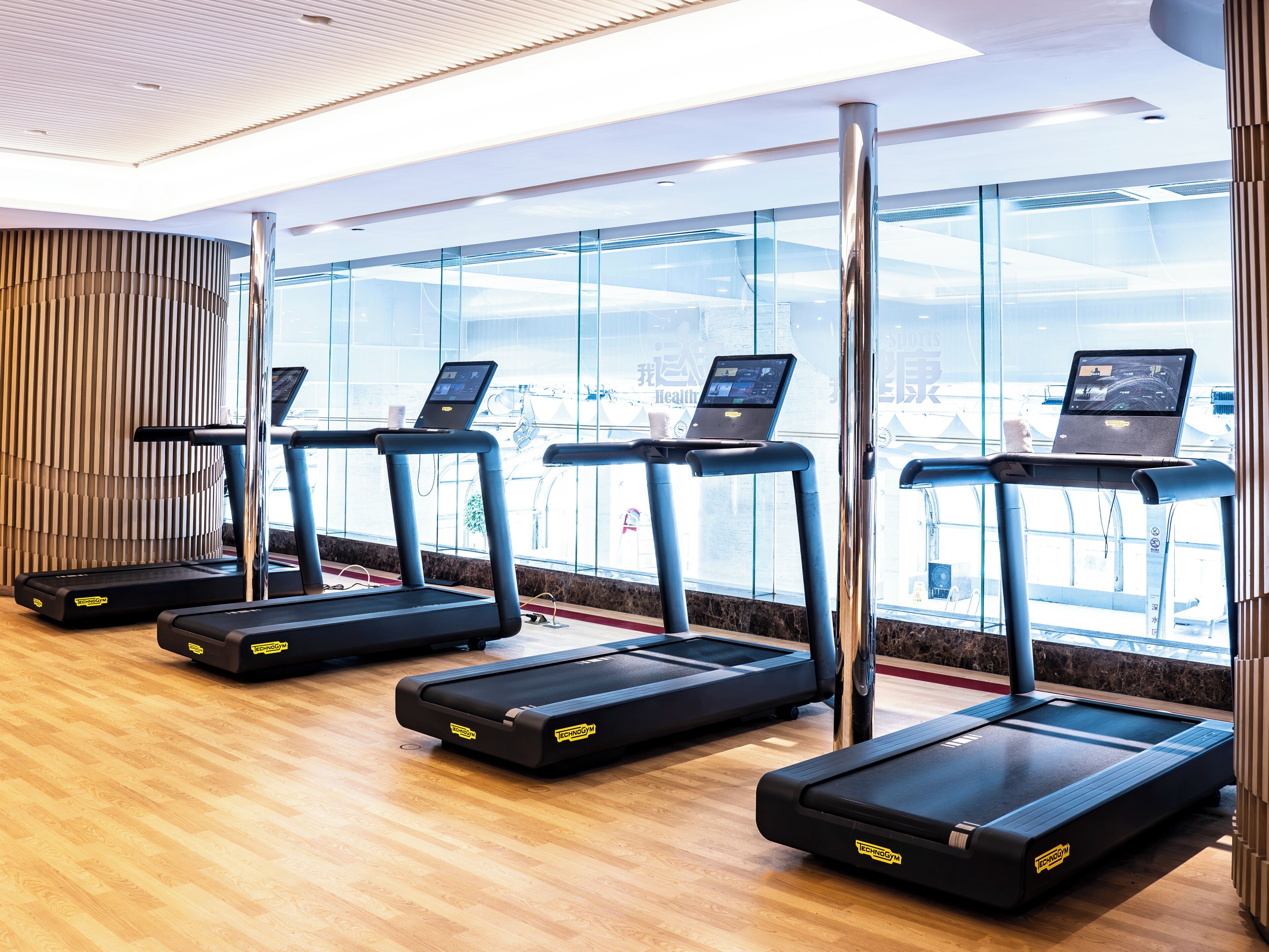 Un gimnasio con cintas de correr, una laptop sobre una de ellas, y un ventanal con vistas.