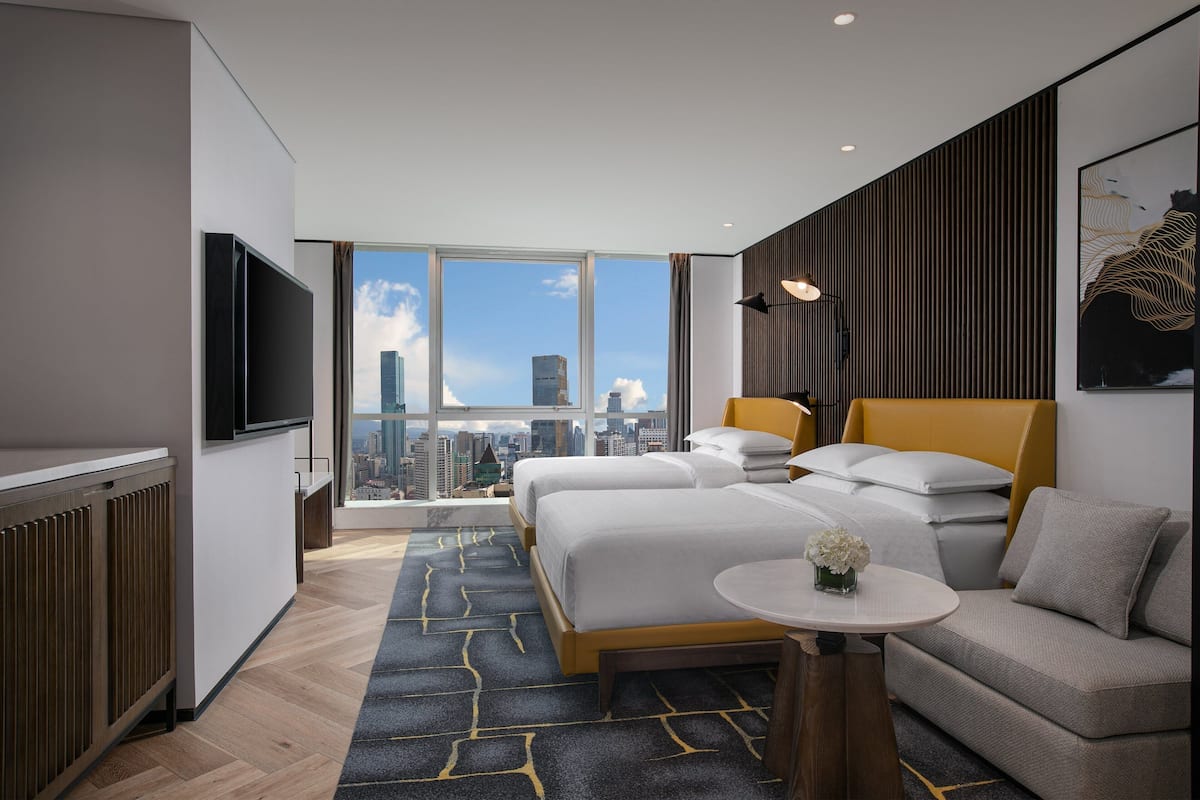 A modern hotel room with two beds, a sofa, a small table, and a TV. The room has a city view through large windows.