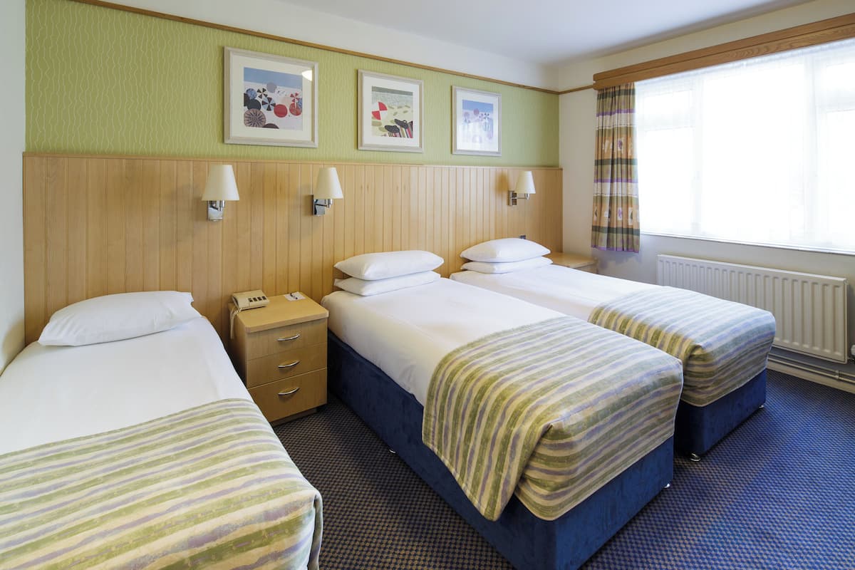 A hotel room with three single beds, a wooden headboard, a small bedside table with a lamp, and a window with curtains.