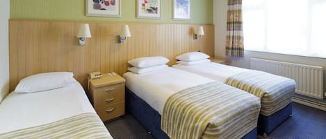 Quarto de hotel com três camas de solteiro, cabeceira de madeira, mesa de cabeceira pequena com abajur e uma janela com cortinas.