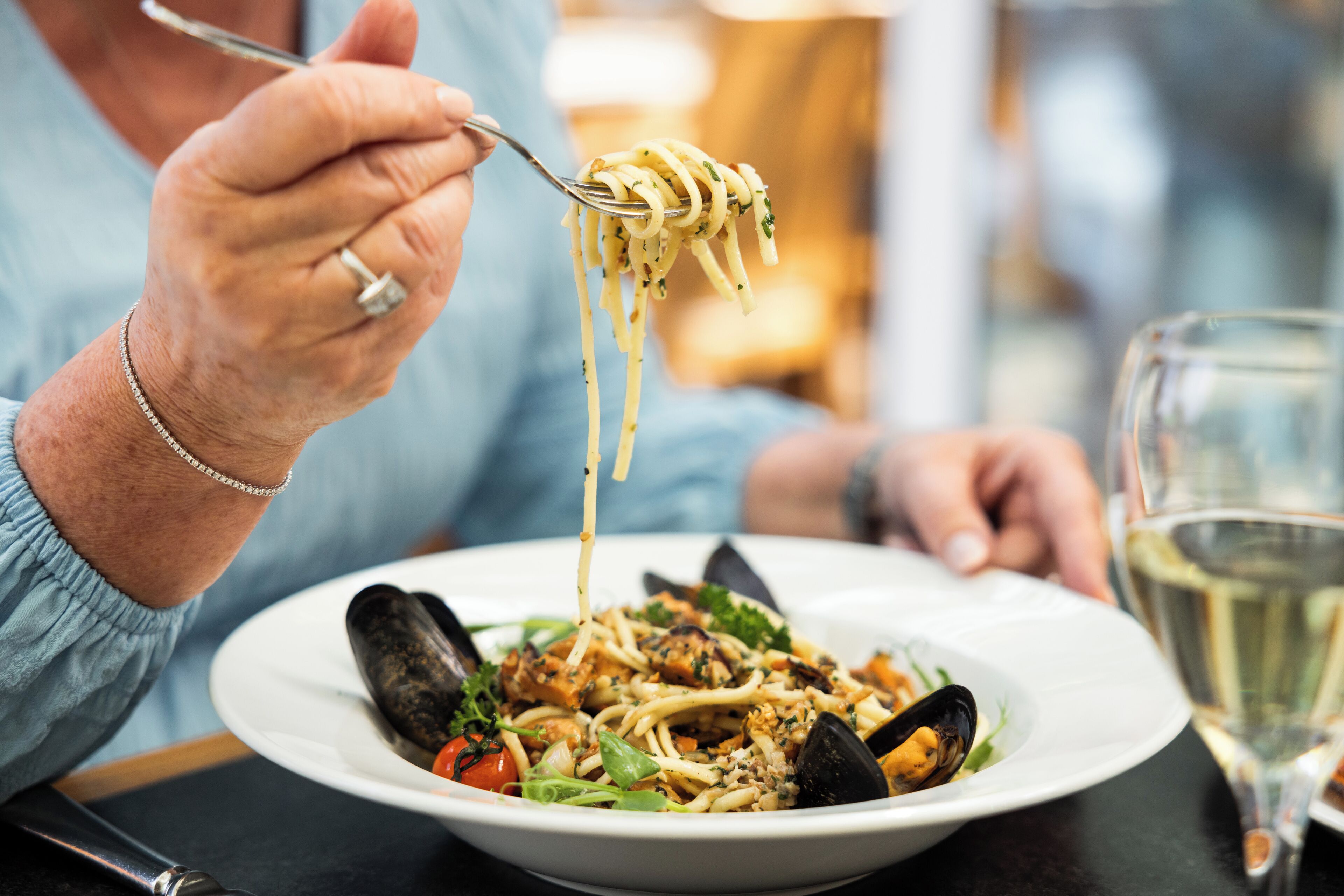 A person holding a fork with spaghetti, a plate with mussels, and a glass of white wine.