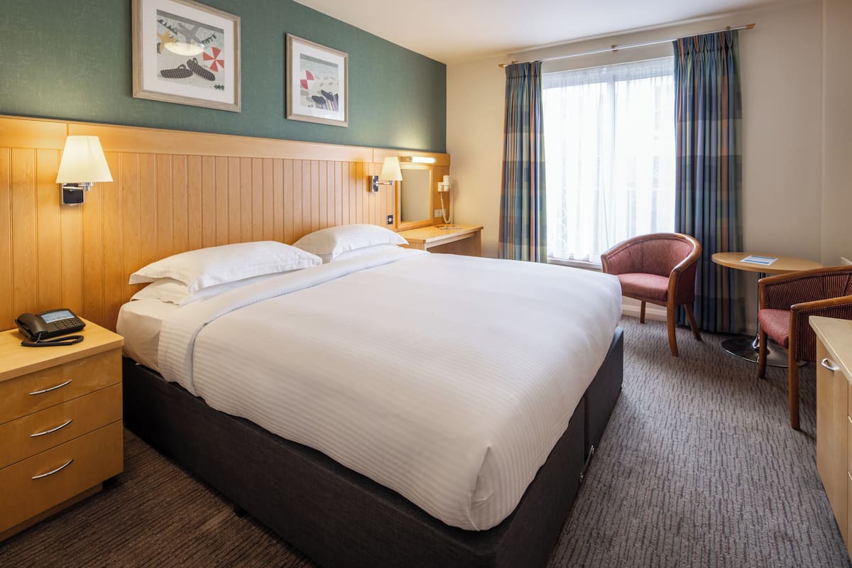 A hotel room with a large bed, bedside tables, a desk, a chair, and a window with curtains.