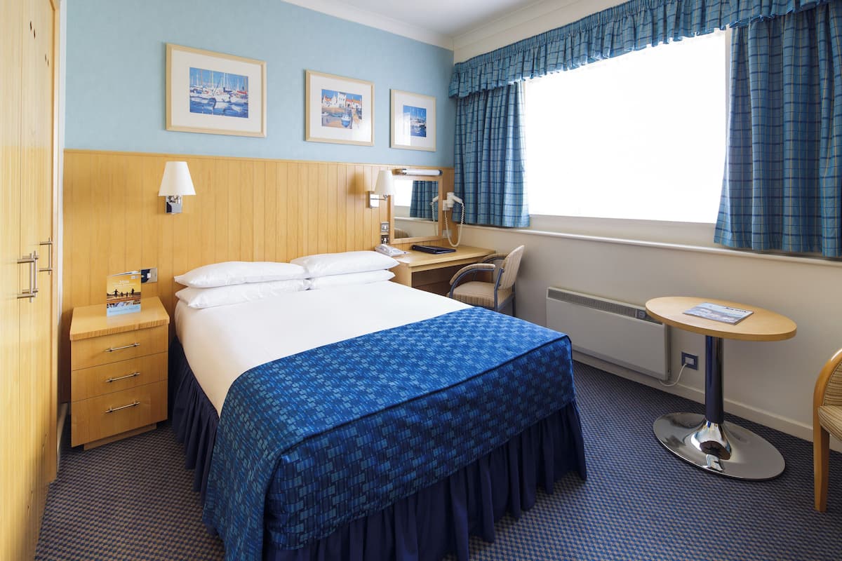 A hotel room with a large bed, a desk with a chair, a small table, and a window with curtains.