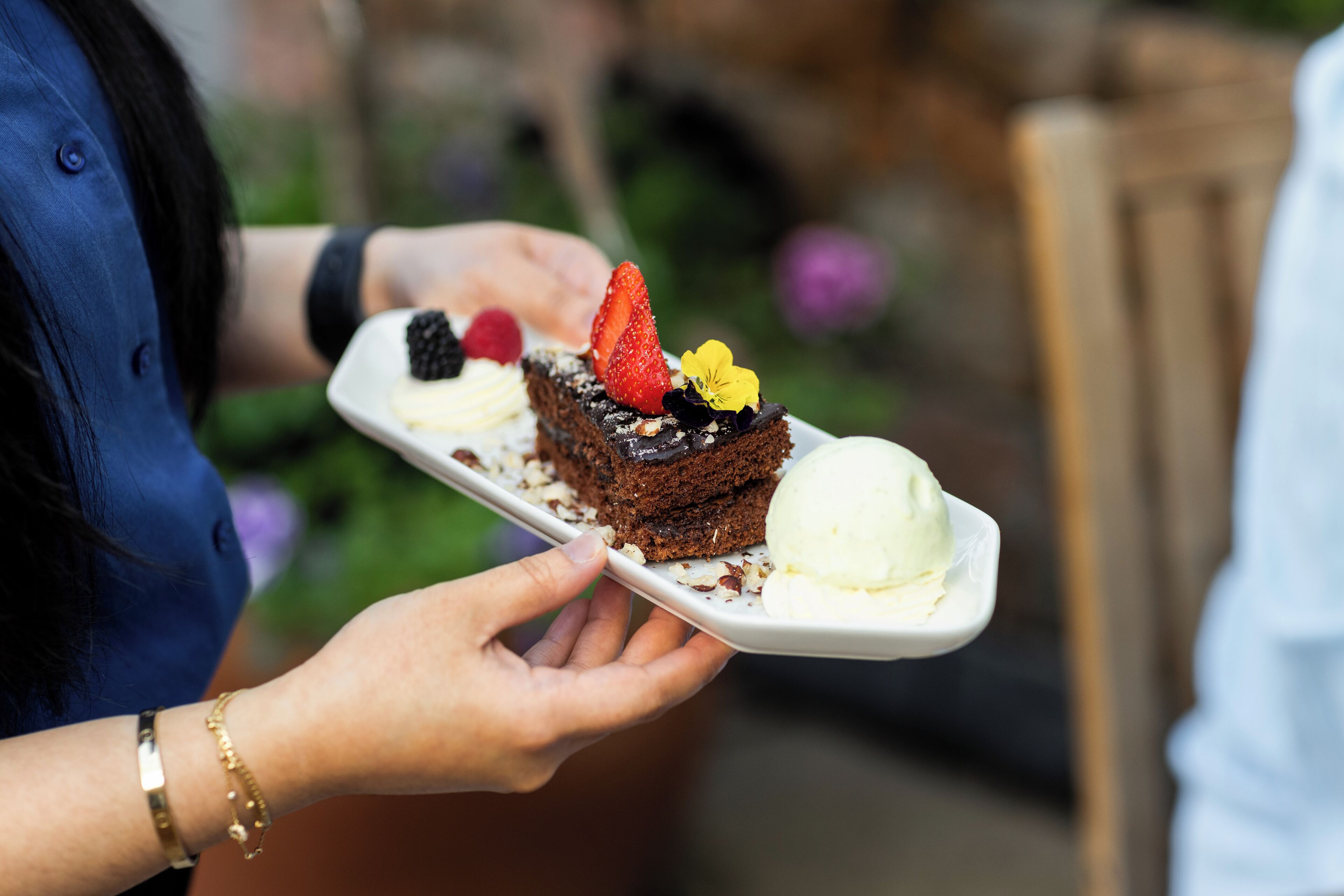 A person holding a white plate with a dessert, including a piece of cake and a scoop of ice cream.