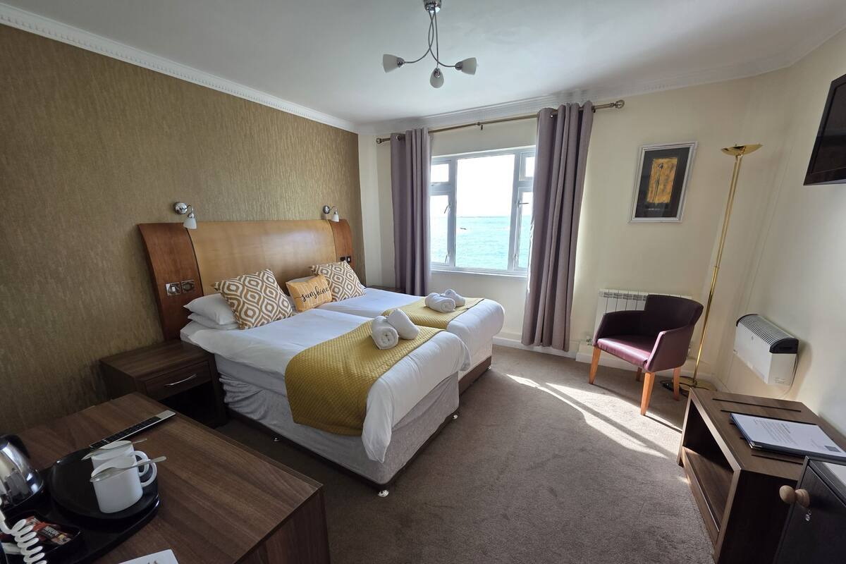 A hotel room with a large bed, a desk with a telephone, a chair, a radiator, and a window with curtains.