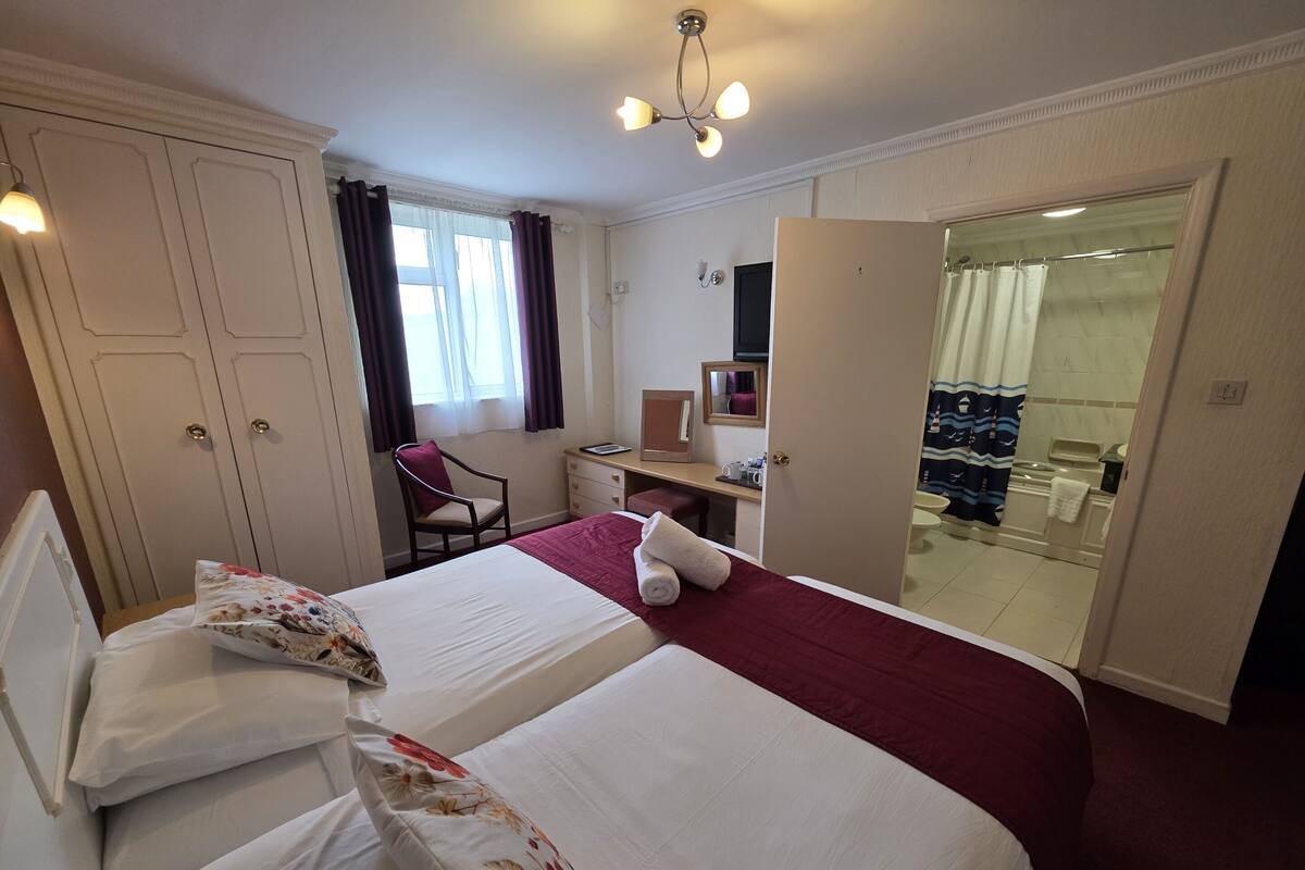 A hotel room with two beds, a desk, a chair, a bathroom visible through an open door, and a ceiling light fixture.