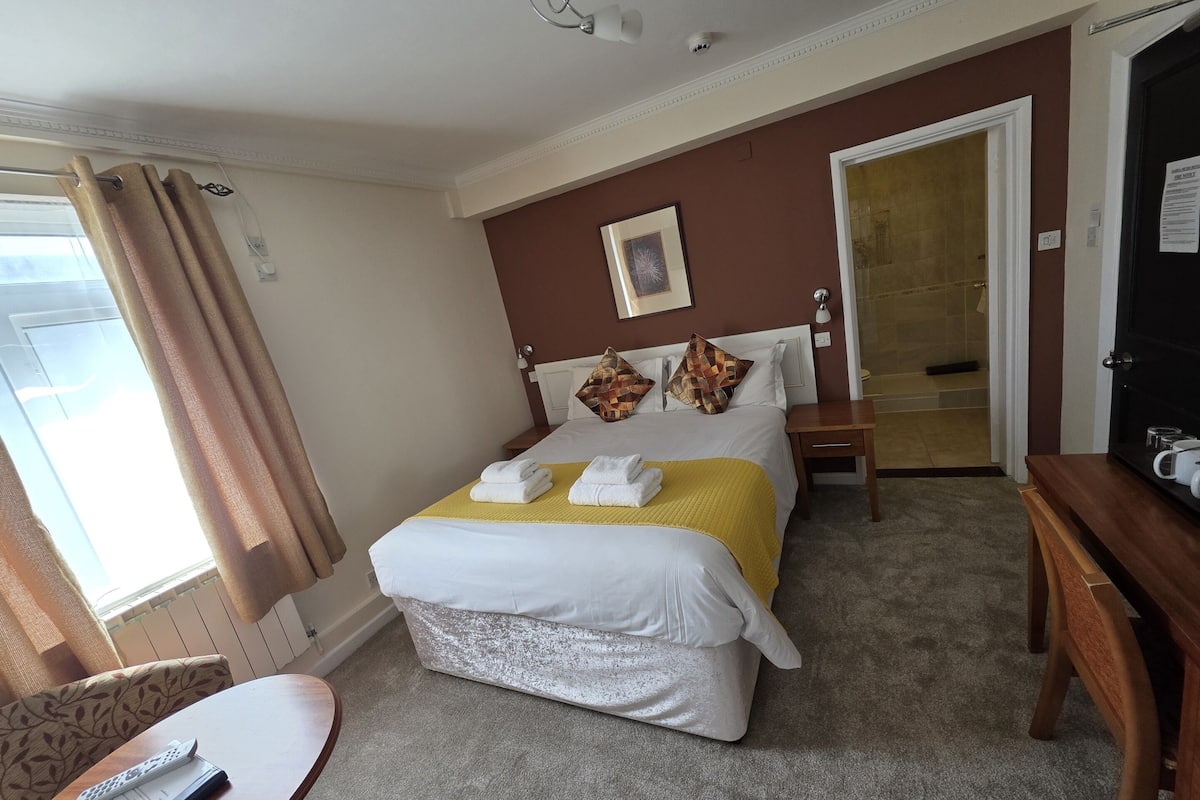 A hotel room with a bed, a bedside table, a desk with a chair, a window with curtains, and a door.