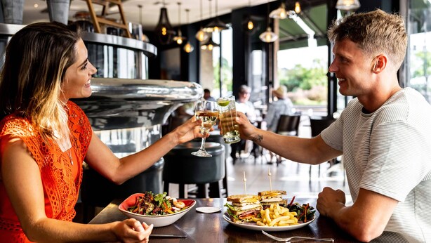 A couple toasting with drinks at a restaurant with a wooden table, plates of food, and a casual atmosphere. - Pomme d'Or Hotel (St. Helier)