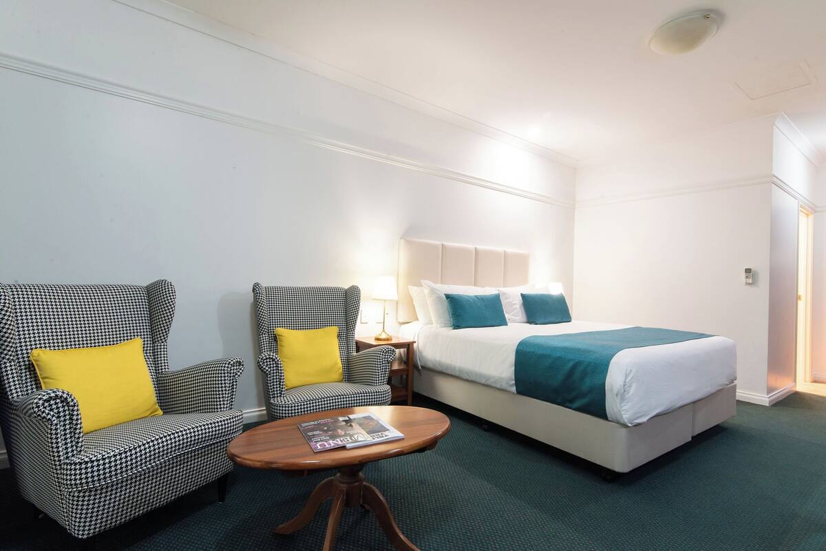Une chambre d’hôtel comprenant un lit, deux fauteuils, une petite table et une table de chevet avec une lampe.