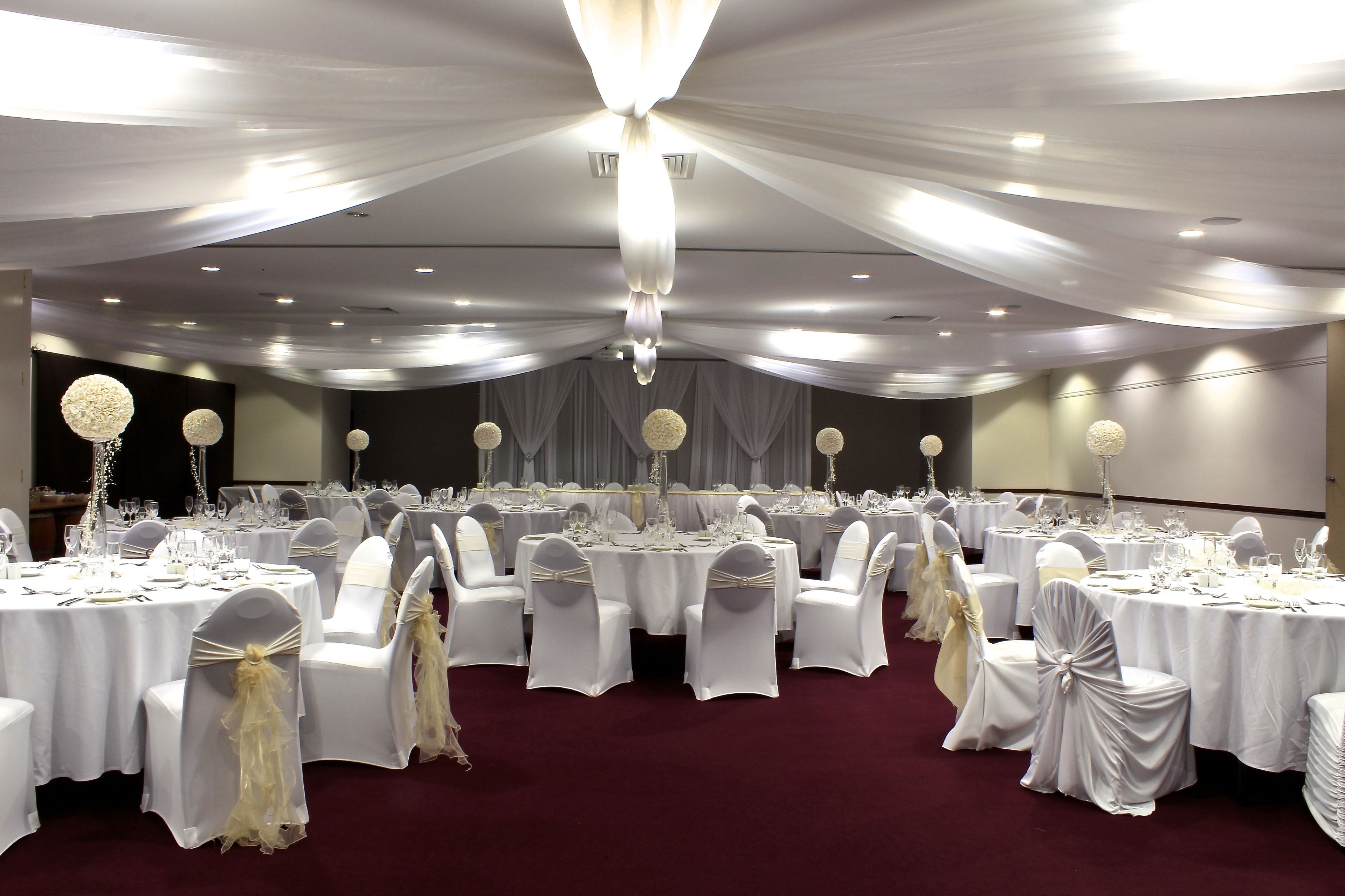 Un salón de banquetes con mesas redondas dispuestas para cenar, sillas blancas con cintas y candelabros.