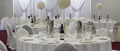 Un salón de banquetes con mesas redondas dispuestas para un evento formal, cada una con mantel blanco, cubiertos y botellas de vino.
