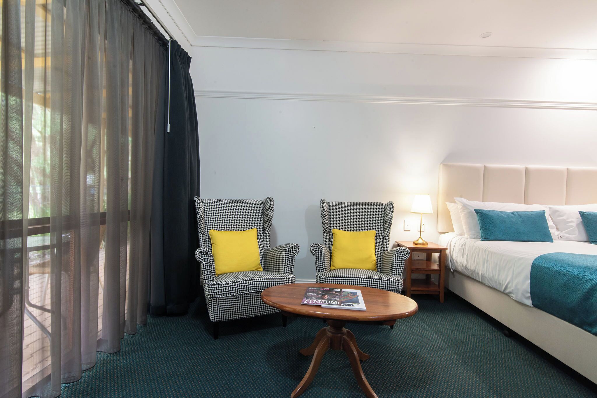 A hotel room with a bed, two armchairs with yellow cushions, a small table, and a window with sheer curtains.