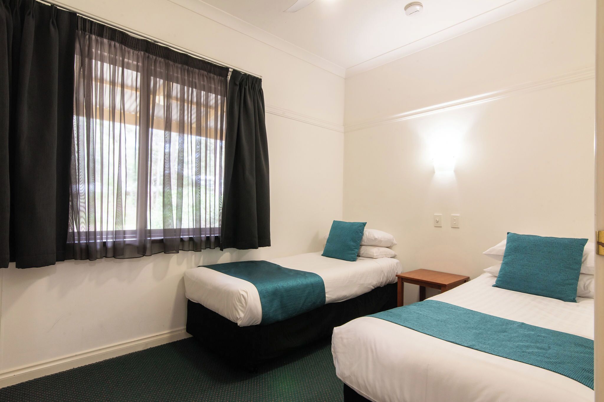 Une chambre d’hôtel avec deux lits, une petite table de chevet, une fenêtre avec des rideaux légers et une lampe fixée au mur.