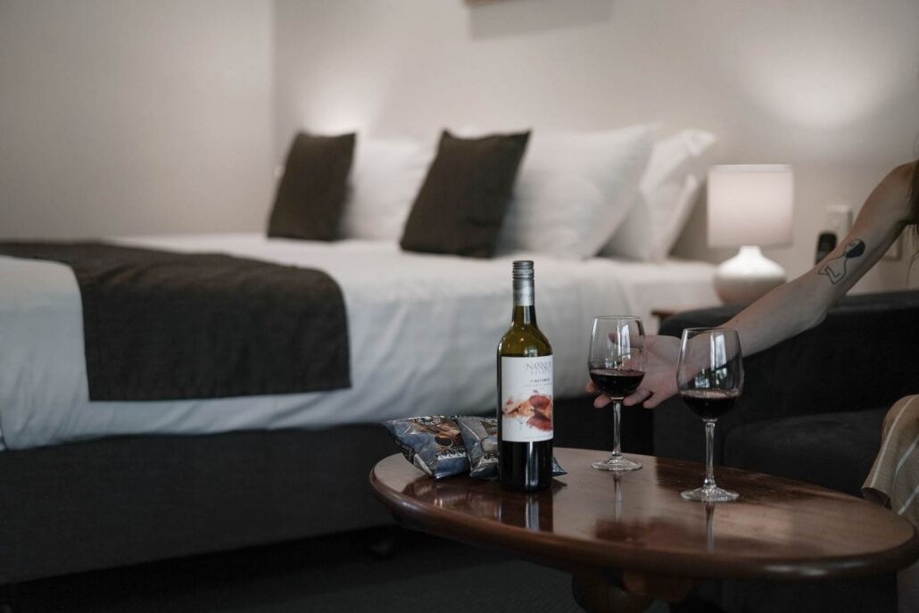 Habitación de hotel con una cama, una mesita de noche con una botella de vino y dos vasos, y una persona con un tatuaje en el brazo.