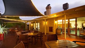 Un espace de restauration en plein air avec des tables et des chaises en bois, un grand parasol et une vue sur l’intérieur du bâtiment.