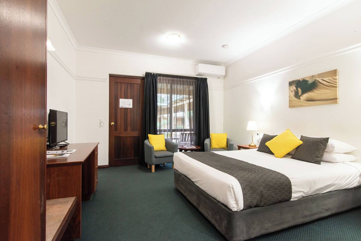Une chambre d’hôtel comprenant un lit, deux fauteuils, un bureau avec un ordinateur, une fenêtre avec des rideaux et un climatiseur fixé au mur.