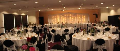 Un salón de banquetes con mesas redondas dispuestas para un evento formal, con manteles blancos, sillas negras y centros de mesa florales.