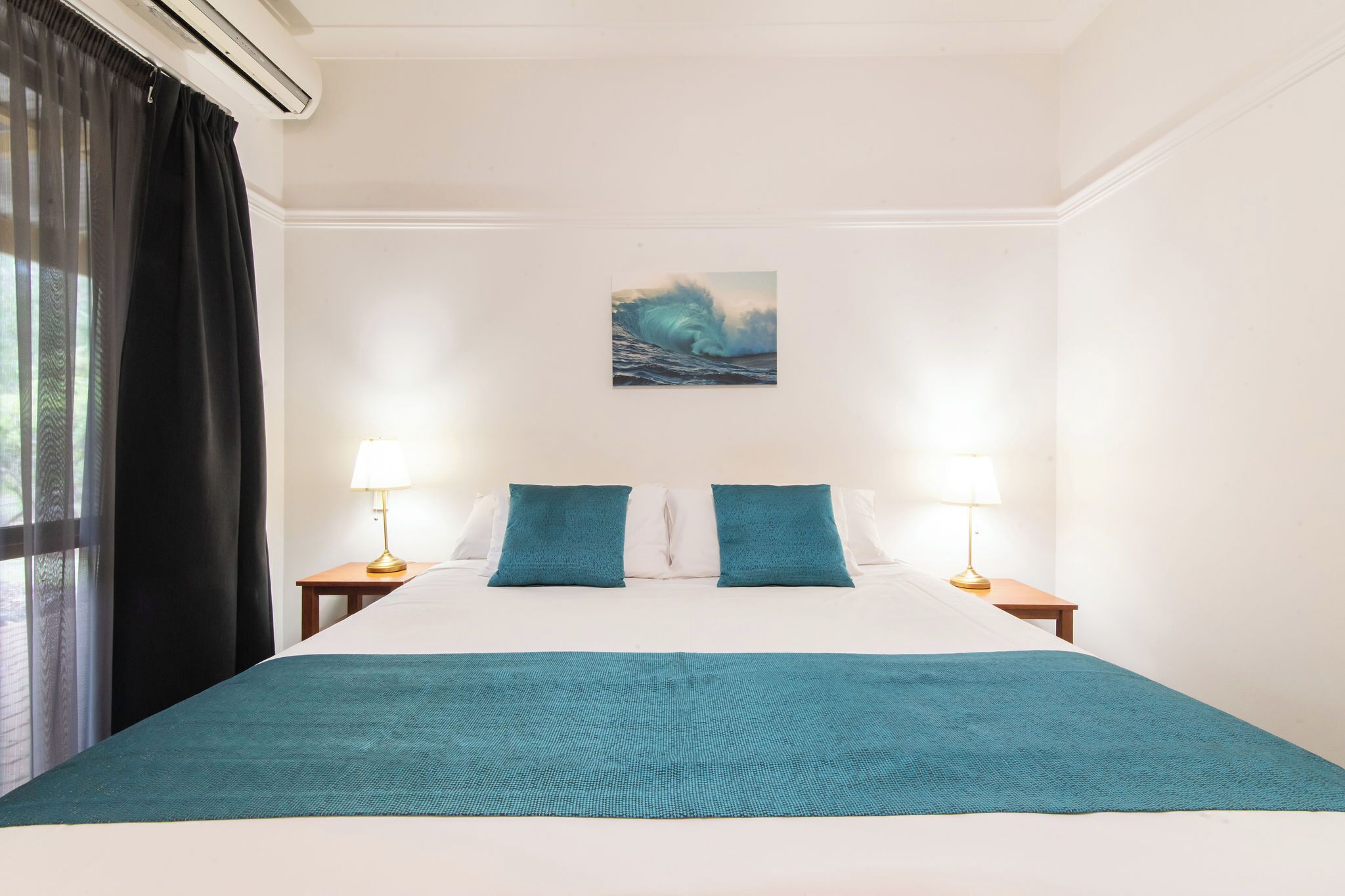 A hotel room with a large bed, two bedside tables with lamps, a window with black curtains, and a painting on the wall.