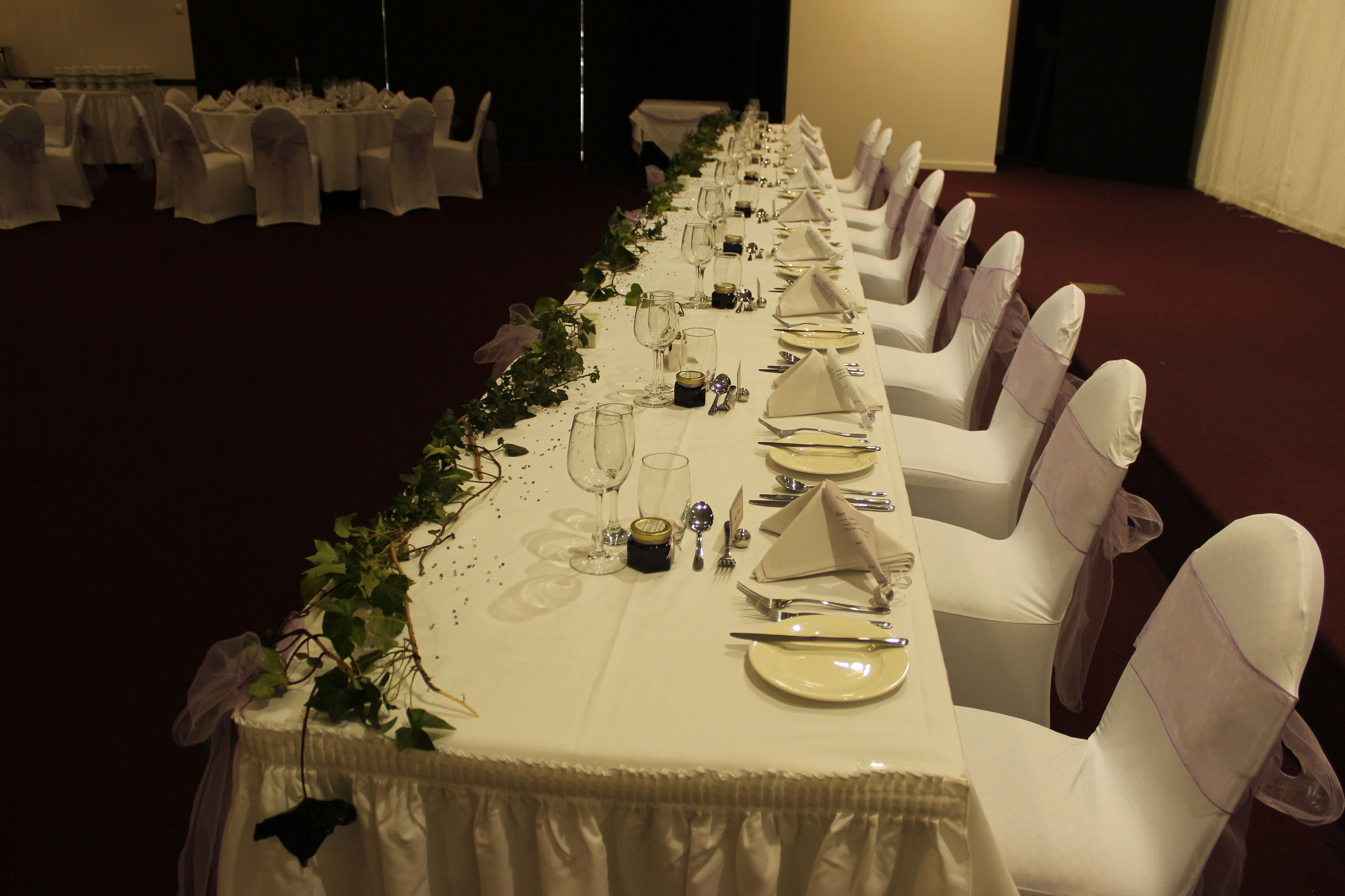A long banquet table set with white tablecloths, silverware, and glassware, surrounded by white chairs with purple sashes.