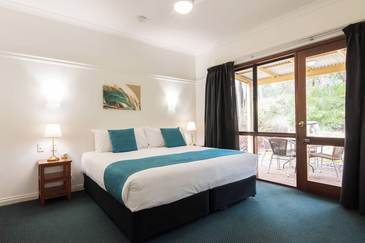 Une chambre d’hôtel avec un grand lit, une table de chevet avec une lampe, un tableau au mur et une porte-fenêtre coulissante donnant sur un espace extérieur aménagé avec des chaises.