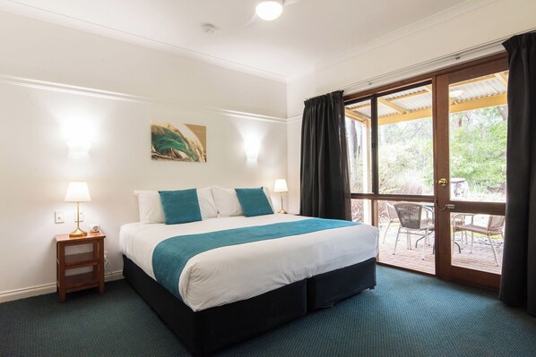 Habitación de hotel con una cama grande, una mesita de noche con lámpara, un cuadro en la pared y un ventanal corredero que da a un espacio exterior con sillas.