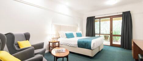 A hotel room with a bed, two armchairs, a small table, a lamp, and a view of the outdoors through a sliding door.