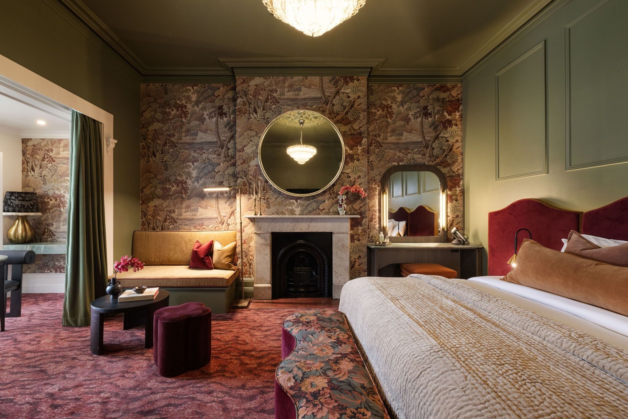 A hotel room with a fireplace, a round mirror, a sofa, a bed, and a small table with a vase.