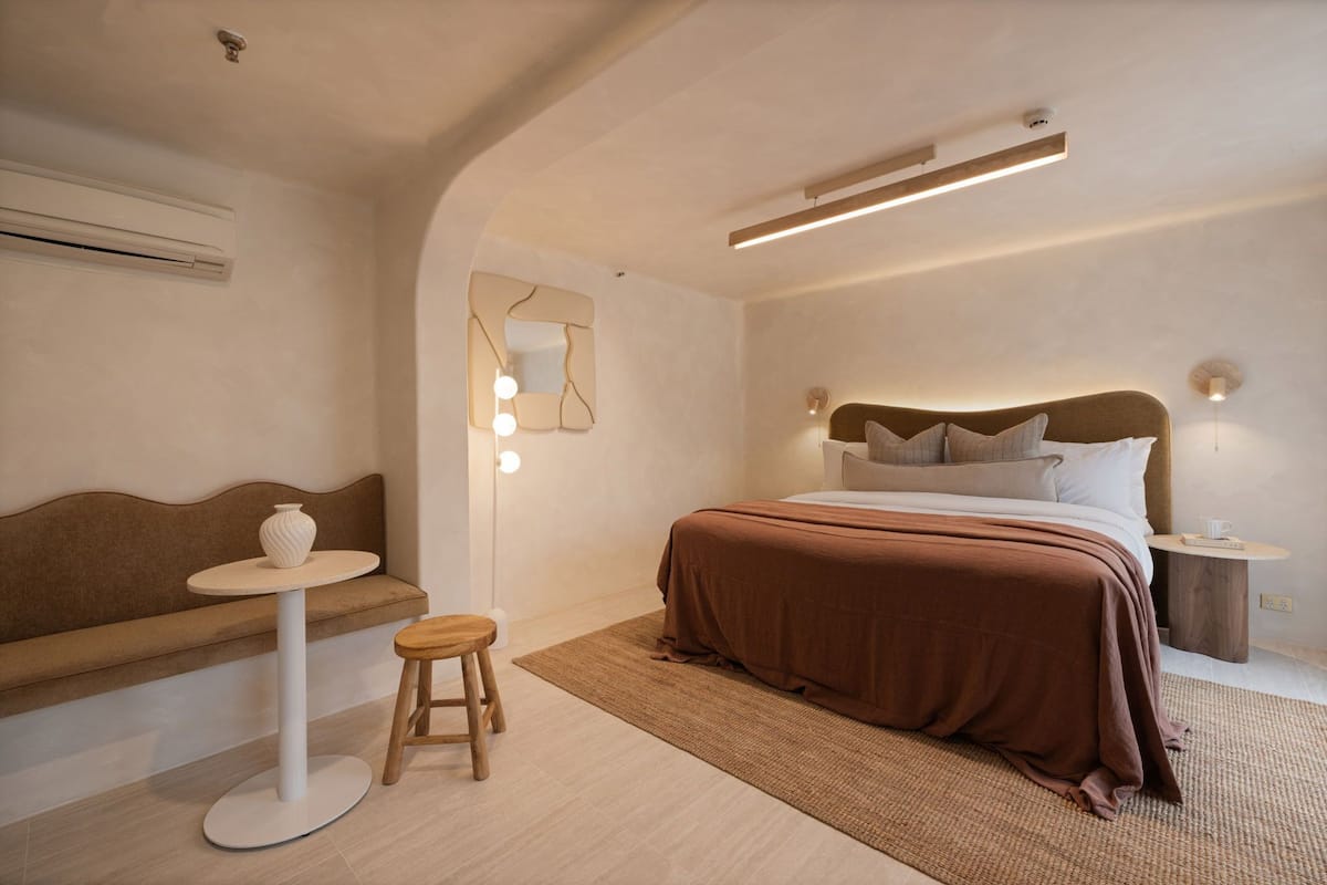 A modern bedroom with a large bed, a small round table, a stool, a bench, and a wall-mounted mirror.