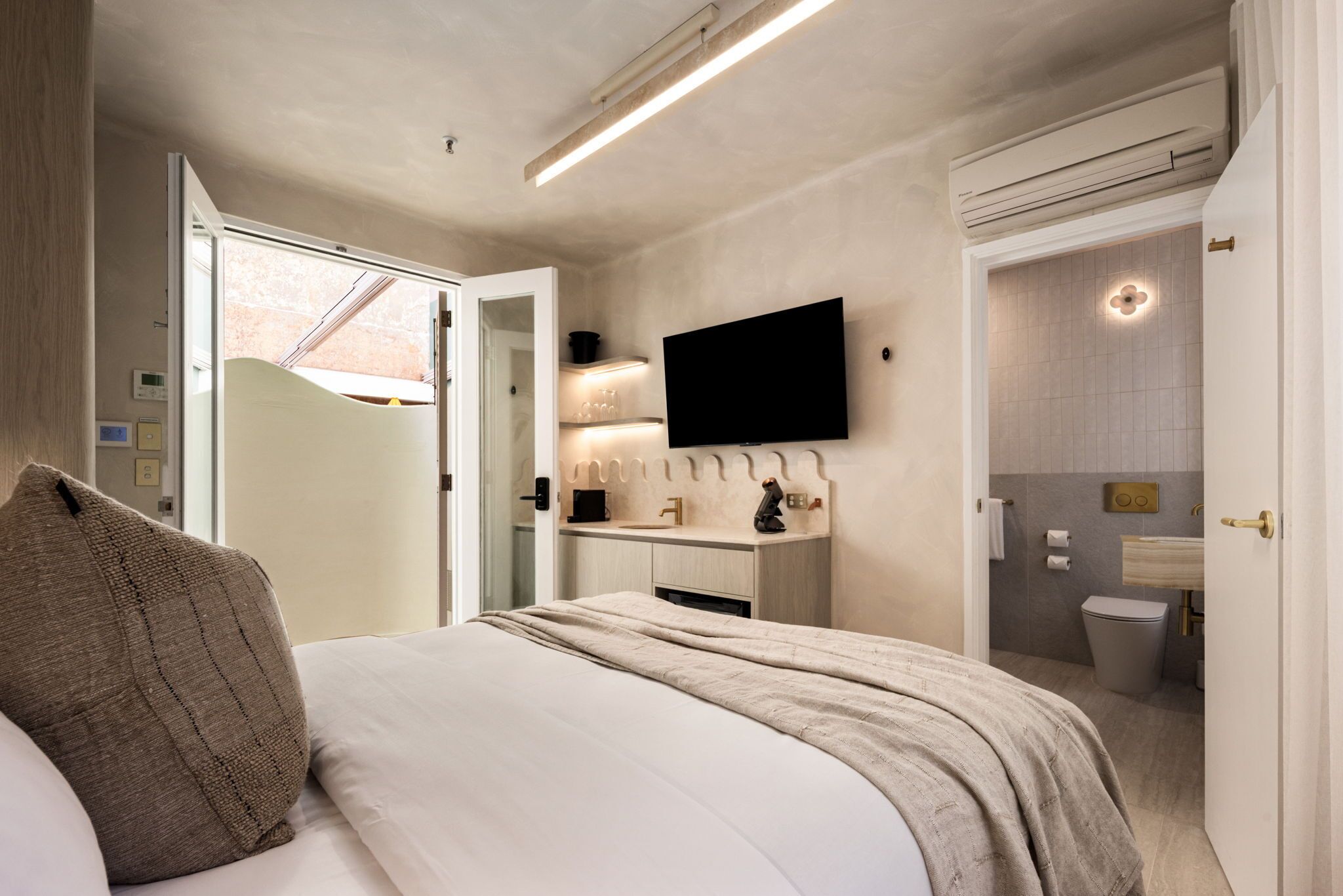 A modern hotel room with a bed, a flat-screen TV mounted on the wall, a bathroom with a toilet and sink, and a window providing natural light.