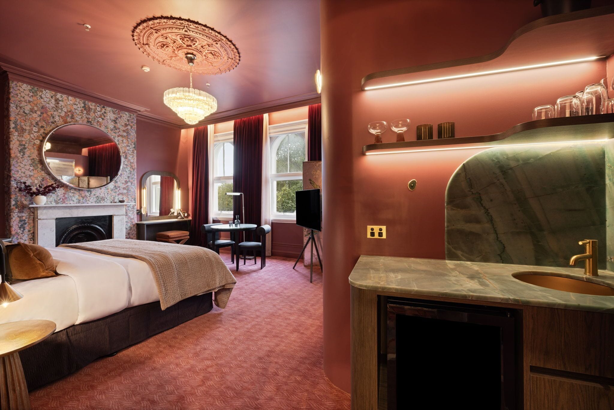 A hotel room with a bed, a desk, a chair, a fireplace, a large mirror, a chandelier, and a marble bathroom vanity.