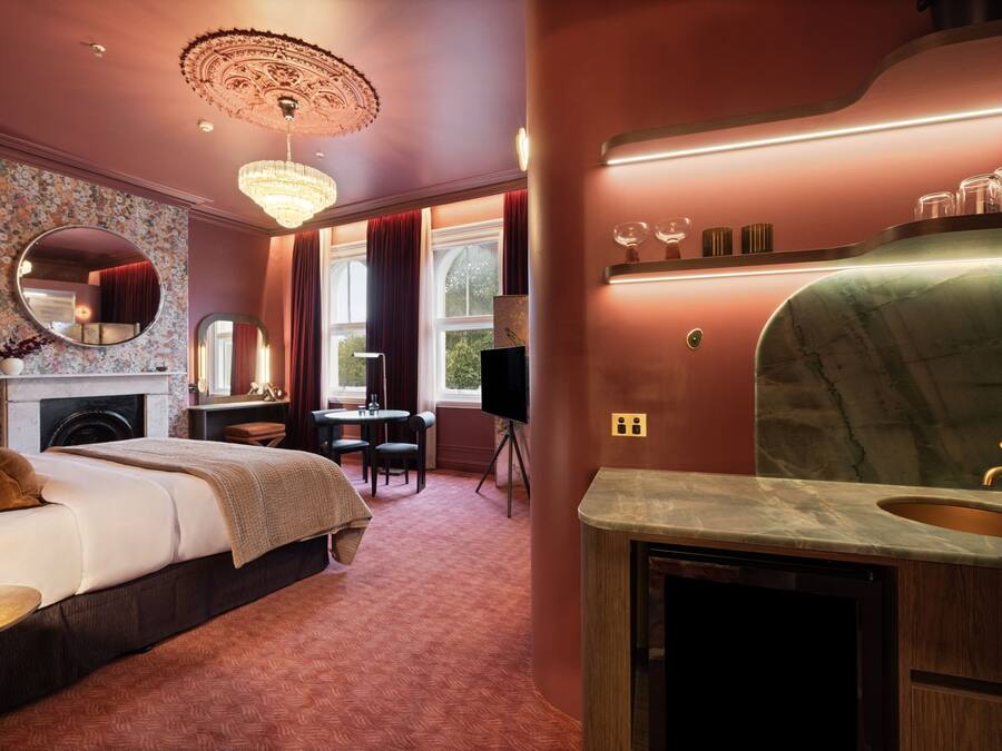 A hotel room with a bed, a desk, a chair, a fireplace, a large mirror, a chandelier, and a marble bathroom vanity.