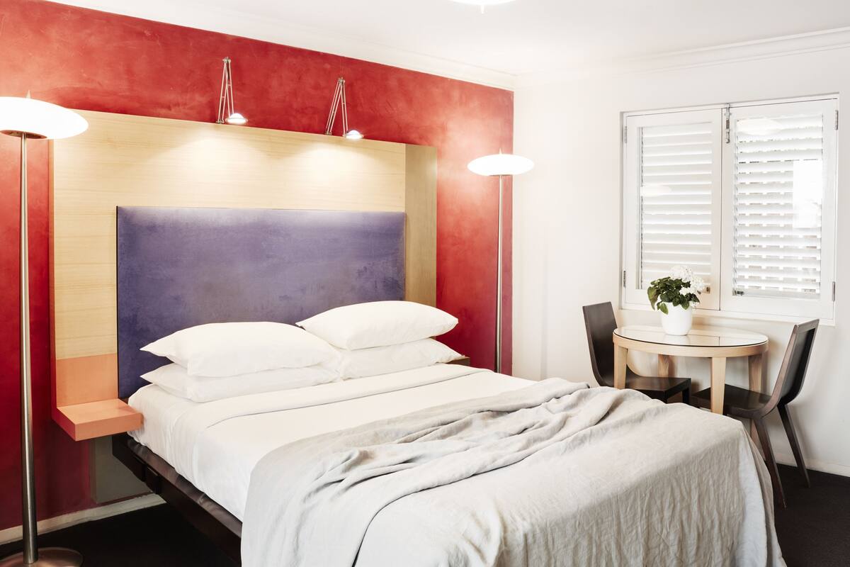 Una habitación de hotel moderna con cama, mesitas de noche, una mesa redonda con sillas y una ventana con persianas.