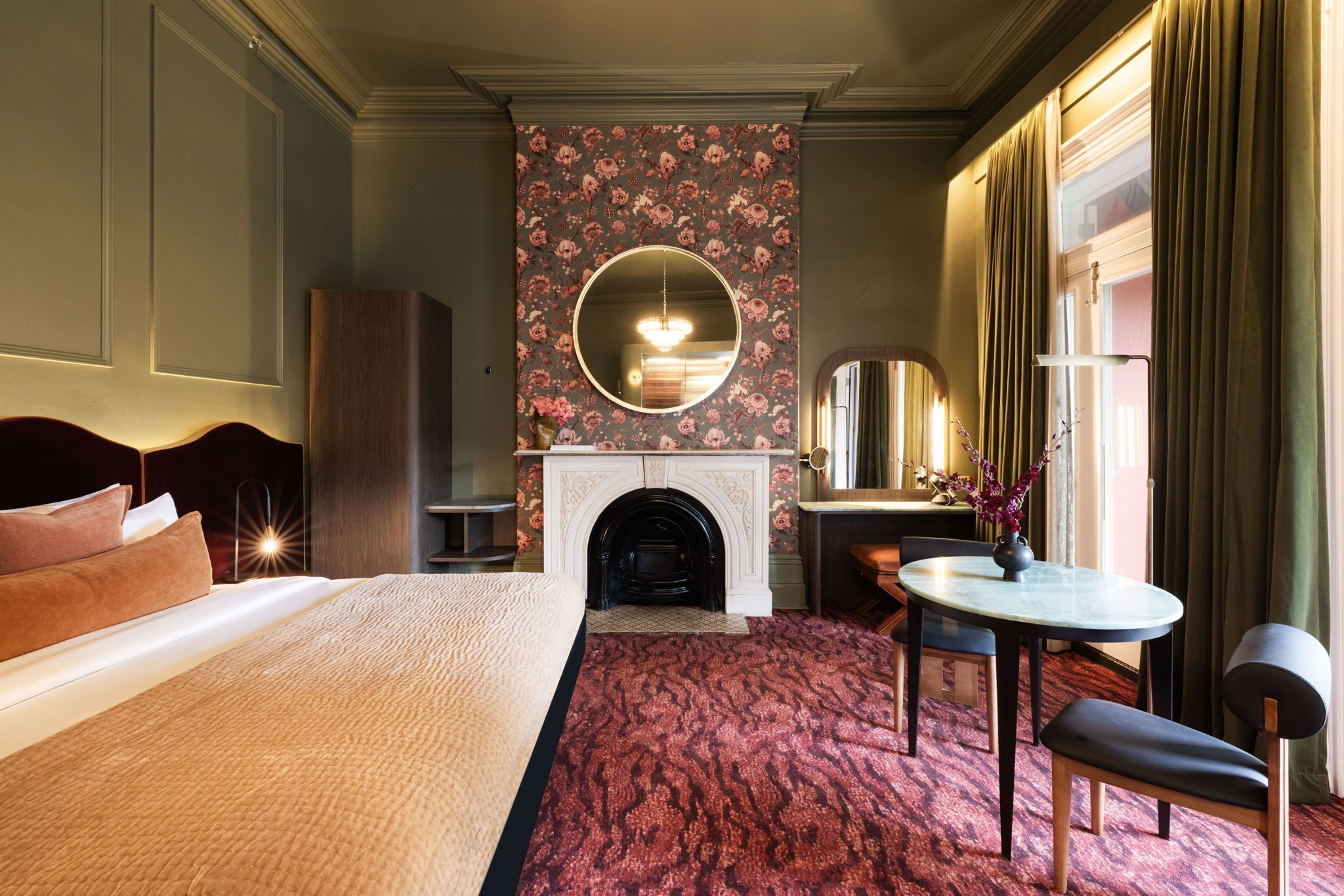 A hotel room with a large bed, a fireplace, a round mirror, a small table, and a chair.