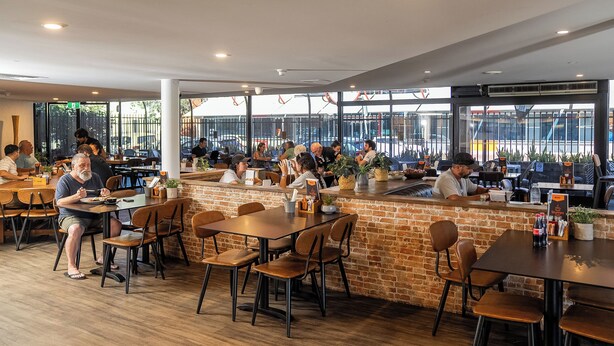 A spacious restaurant with multiple tables and chairs, a brick accent wall, and large windows offering views of the outside.
