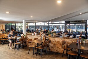 A spacious restaurant with multiple tables and chairs, a brick accent wall, and large windows offering views of the outside. - Greet Hotel Alice Springs (Alice Springs)