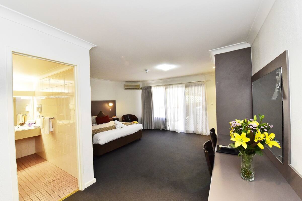 A hotel room with a bed, a desk, a chair, a bathroom with a shower, and a vase with flowers.