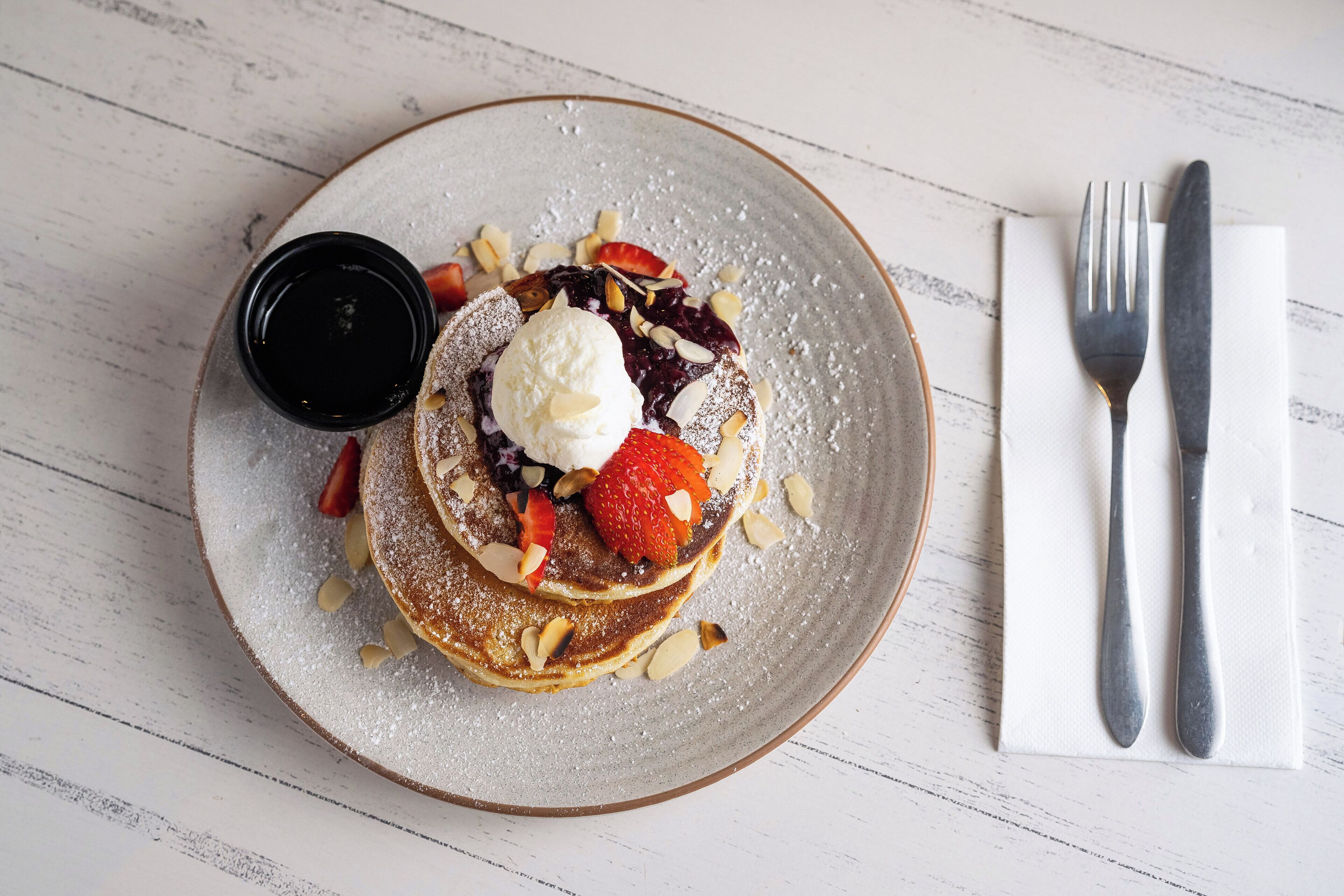 A plate of pancakes with strawberries, a dollop of cream, and a small container of syrup.