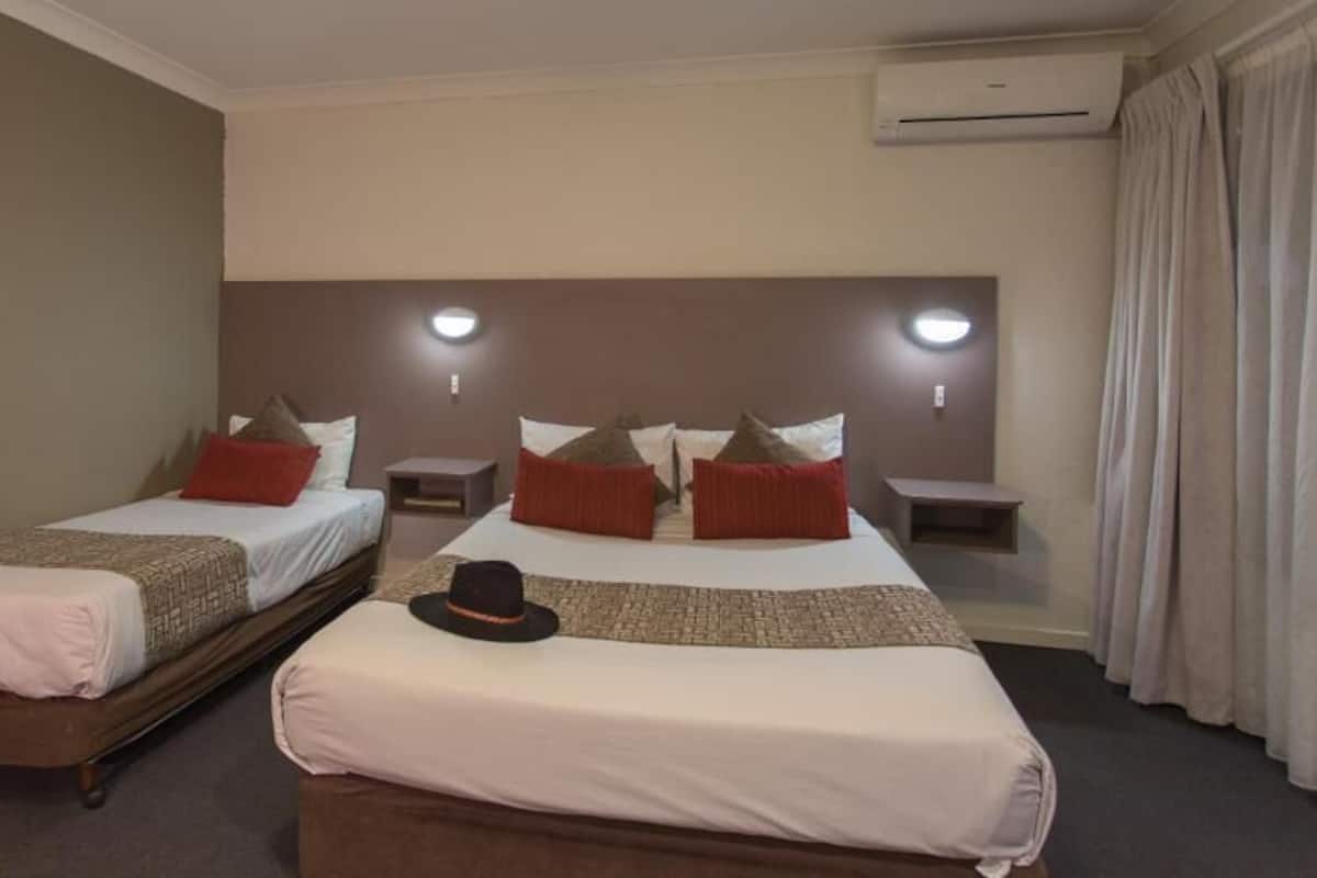 Ein Hotelzimmer mit zwei Einzelbetten, einem großen Bett, einem Nachttisch, einer Klimaanlage und einem Fenster mit Vorhängen.