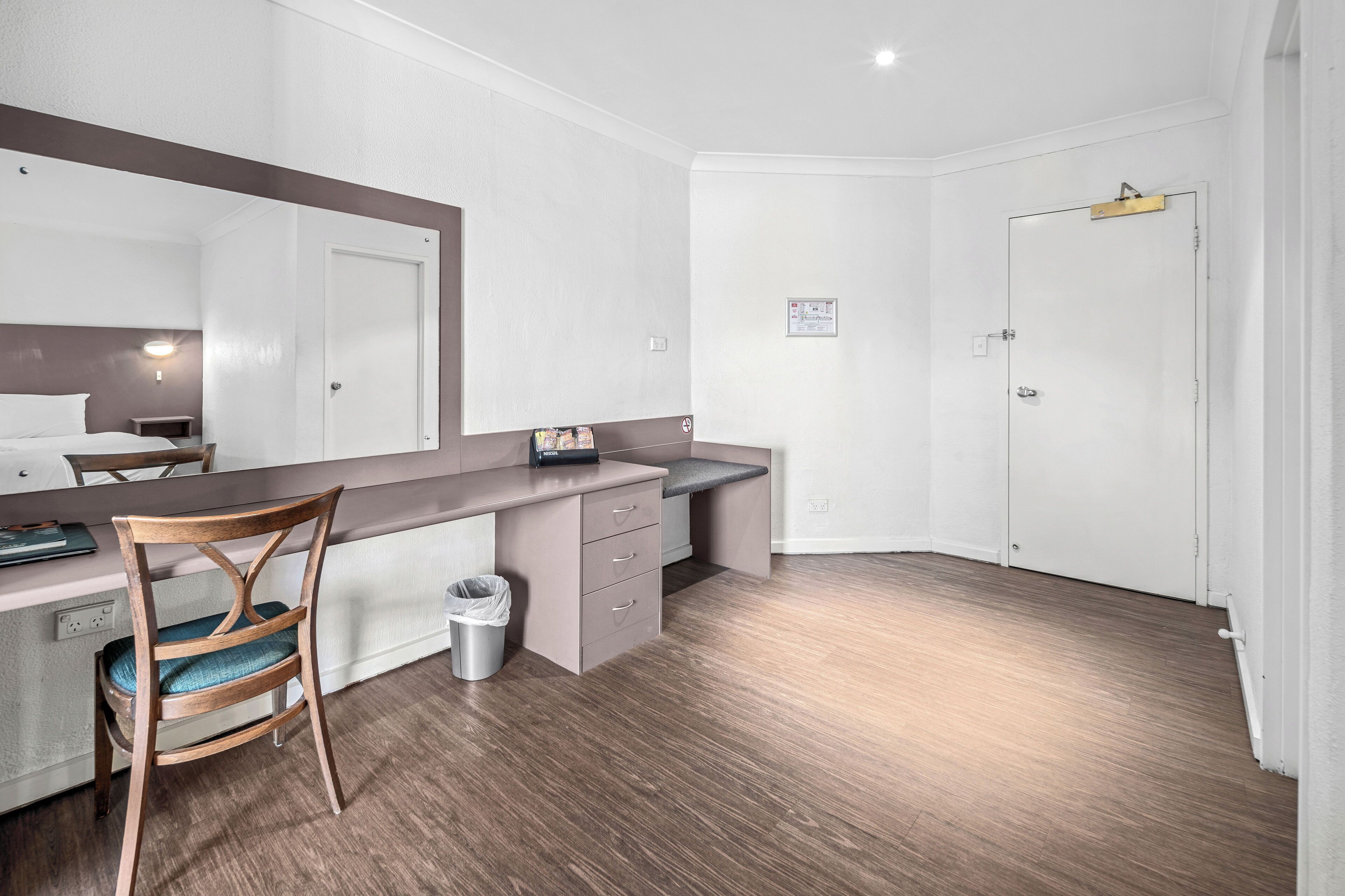 Una habitación de hotel moderna con un escritorio de madera, una silla, un pequeño cubo de basura y una puerta blanca.