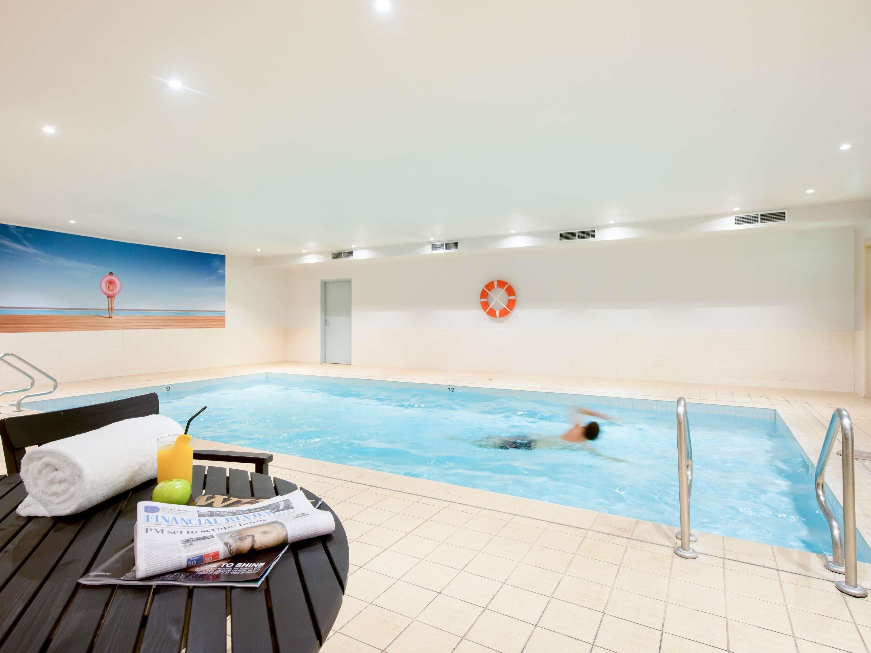 An indoor swimming pool with a lifebuoy on the wall, a towel, a glass of orange juice, and a newspaper on a table.