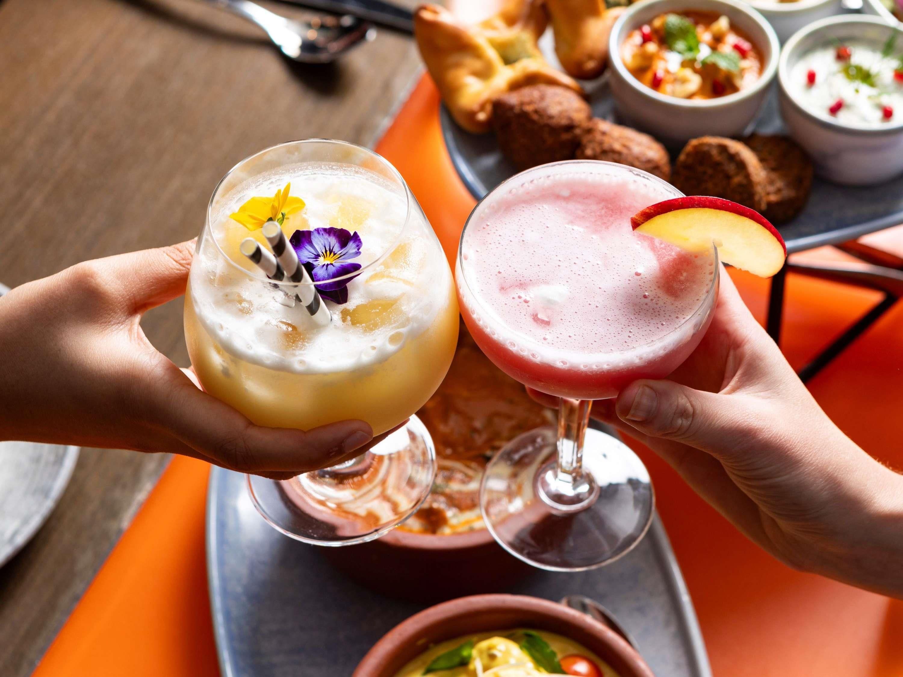 Dos personas sosteniendo bebidas, con una bandeja de comida y bebidas al fondo.