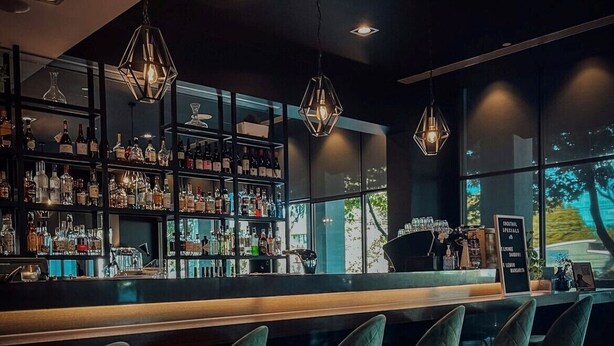 Un'area bar con un lungo bancone, numerose bottiglie sugli scaffali e lampade a sospensione.