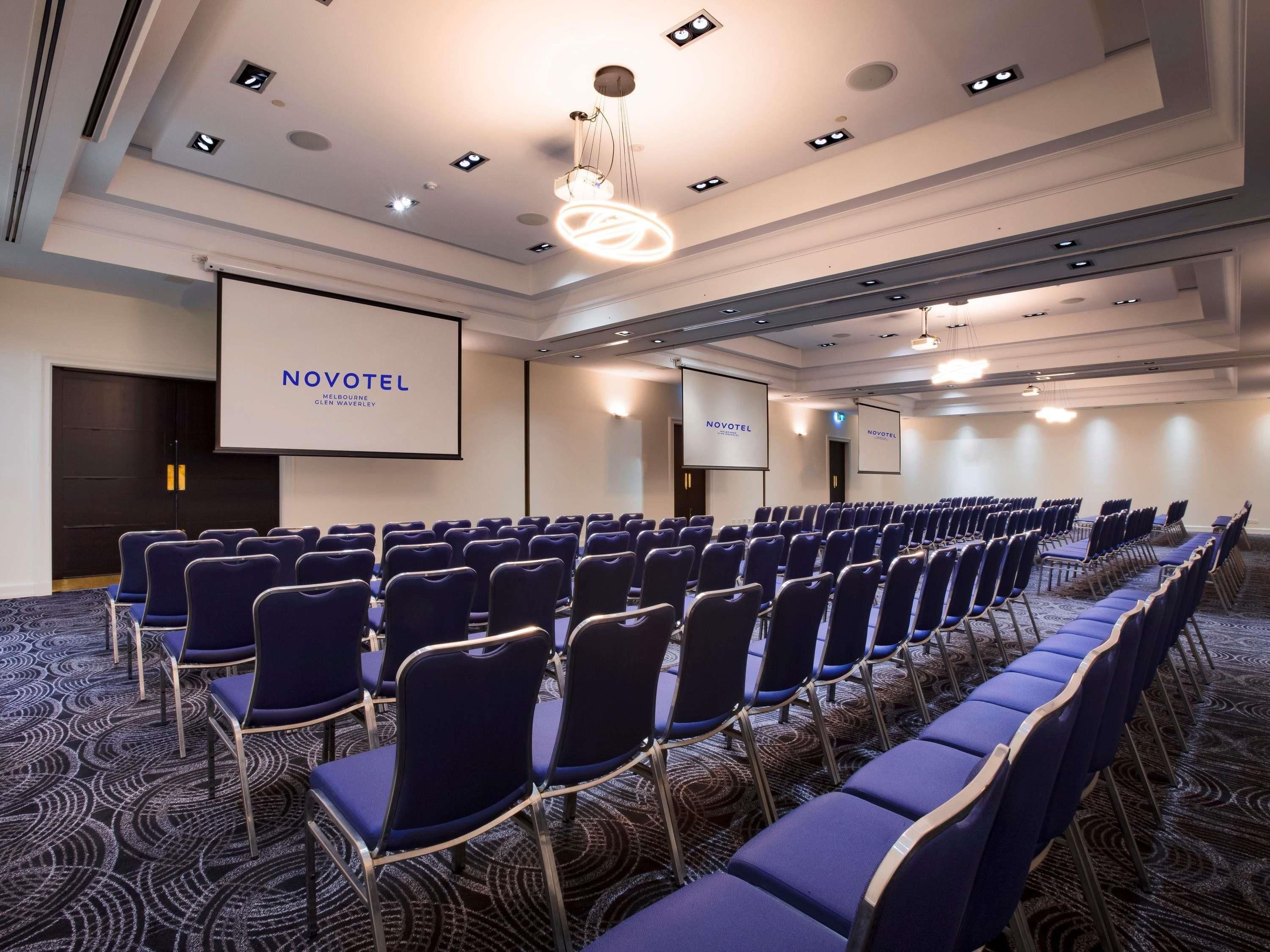 Uma sala de conferências com fileiras de cadeiras, uma tela grande e piso coberto de carpete.