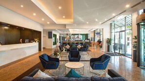 Hall d'albergo con reception, area salotto con divani e poltrone, un bar e ampie finestre.