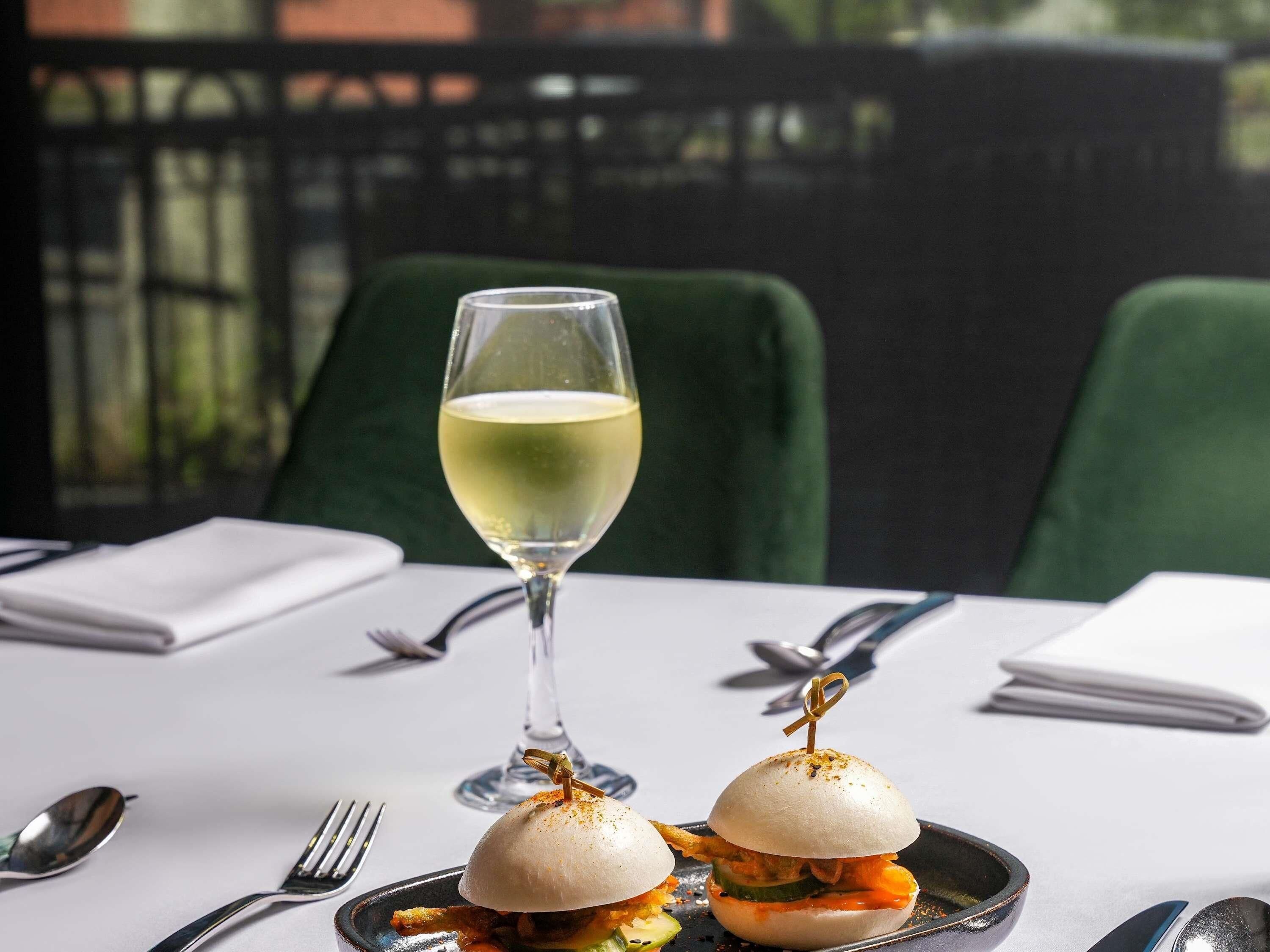 A table with a glass of white wine, two sandwiches, and dining utensils.