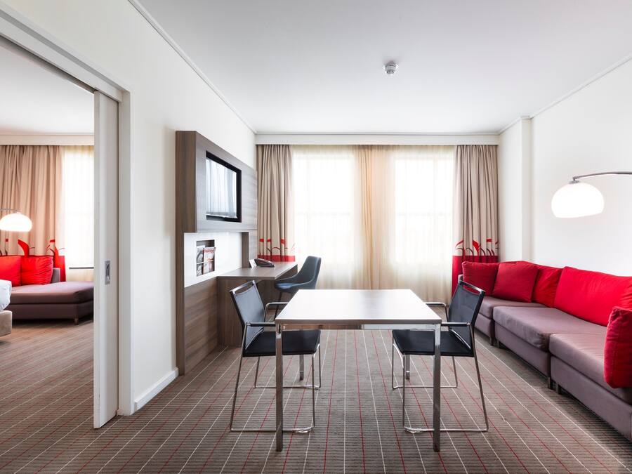 A modern hotel room with a dining table, two chairs, a sofa, a TV, and a bookshelf.