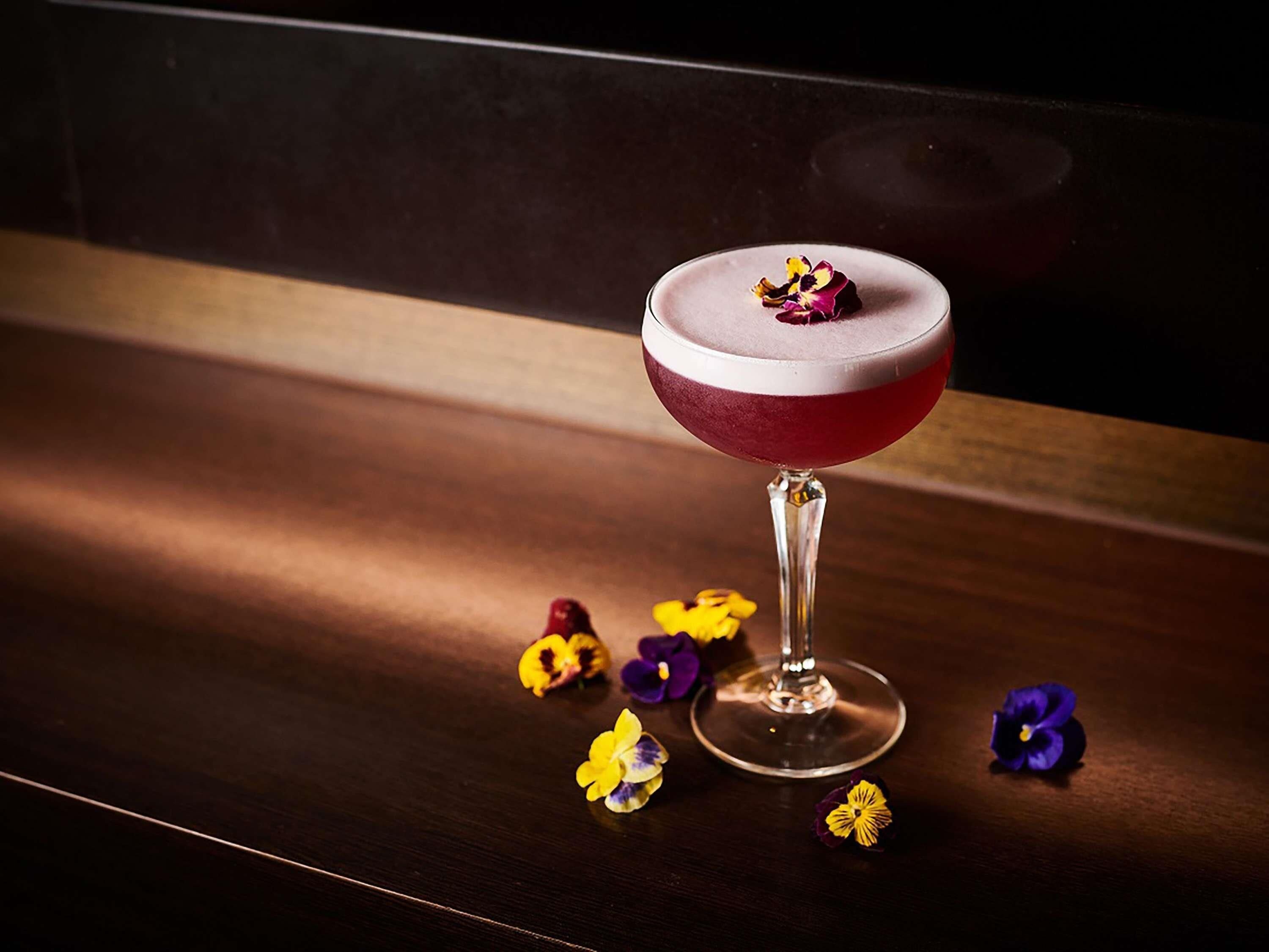 A cocktail in a stemmed glass on a wooden surface with scattered flowers.